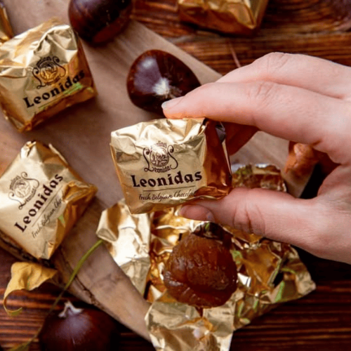 Hand holding a Leonidas chocolate truffle surrounded by more truffles and packaging on a wooden surface.